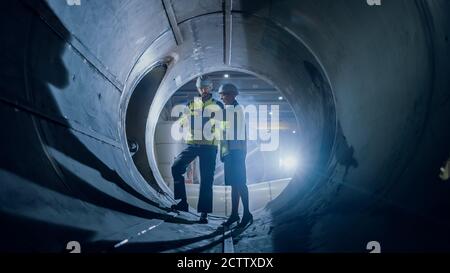 Two Heavy Industry Engineers Walking Inside Pipe, Use Laptop, Have Discussion, Checking Design. Construction of the Oil, Natural Gas and Biofuels Stock Photo
