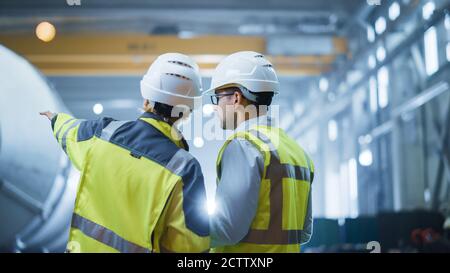 Two Heavy Industry Engineers Stand in Pipe Manufacturing Factory, Use Digital Tablet Computer, Have Discussion. Construction of Oil, Gas and Fuels Stock Photo