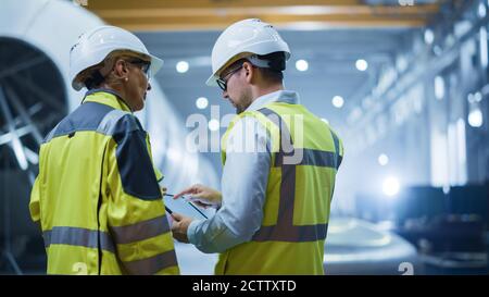 Two Heavy Industry Engineers Stand in Pipe Manufacturing Factory, Use Digital Tablet Computer, Have Discussion. Construction of Oil, Gas and Fuels Stock Photo