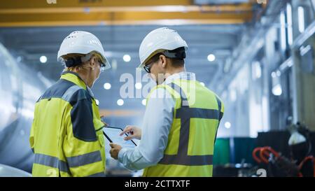 Two Heavy Industry Engineers Stand in Pipe Manufacturing Factory, Use Digital Tablet Computer, Have Discussion. Large Pipe Assembled. Design and Stock Photo