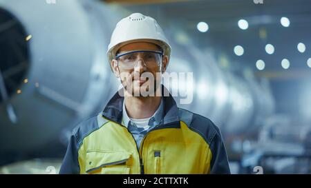 Portrait of Smiling Professional Heavy Industry Engineer Worker Wearing Safety Uniform, Goggles and Hard Hat. In the Background Unfocused Large Stock Photo
