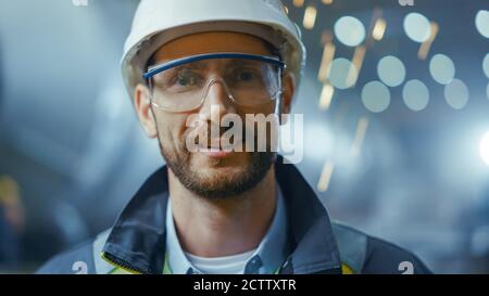 Portrait of Professional Heavy Industry Engineer Worker Wearing Safety Uniform, Goggles and Hard Hat Smiling. In the Background Unfocused Large Stock Photo