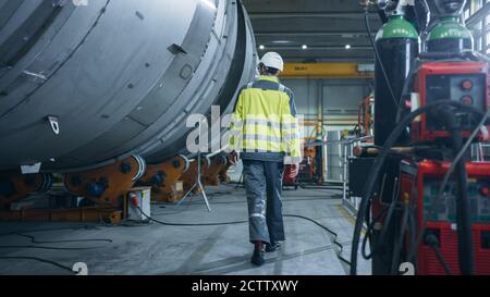 Following Shot of Heavy Industry Engineer Walking through Pipe Manufacturing Factory. Modern Facility for Design and Construction of Large Diameter Stock Photo