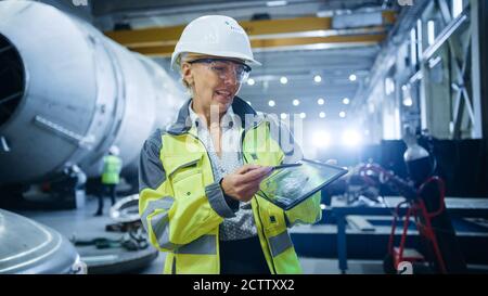 Professional Heavy Industry Female Engineer Wearing Safety Uniform, Holds Digital Tablet Computer and Explains Product Design. Industrial Factory Stock Photo