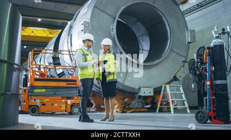 Two Heavy Industry Engineers Stand in Pipe Manufacturing Factory, Use Digital Tablet Computer, Have Discussion. Facility for Construction of Oil, Gas Stock Photo