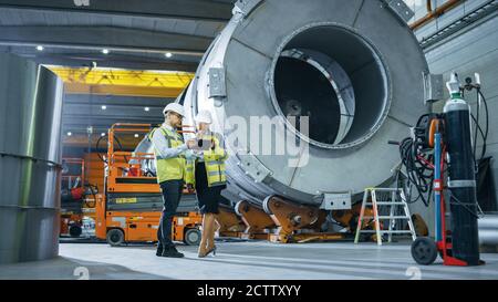 Two Heavy Industry Engineers Stand in Pipe Manufacturing Factory, Use Digital Tablet Computer, Have Discussion. Facility for Construction of Oil, Gas Stock Photo
