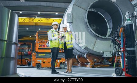 Two Heavy Industry Engineers Stand in Pipe Manufacturing Factory, Use Digital Tablet Computer, Have Discussion. Facility for Construction of Oil, Gas Stock Photo