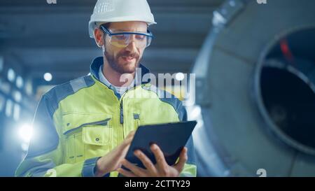 Portrait of Professional Heavy Industry Engineer Worker Wearing Safety Uniform and Hard Hat Uses Tablet Computer. In the Background Construction Stock Photo