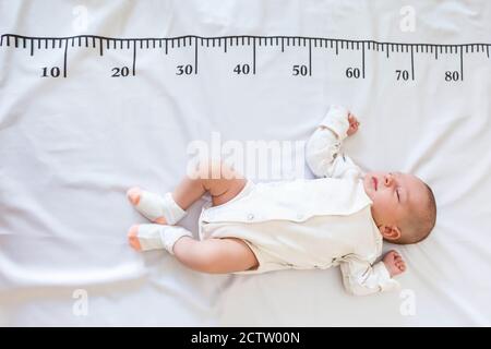 A newborn 2 month baby in white sleeps on a bed on which a measuring ruler for growth is drawn Stock Photo