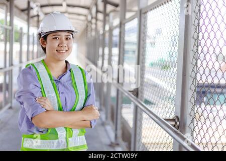Young asian business engineer or technician inspector woman in safety helmet standing arms crossed at construction work site smiling looking camera. Stock Photo
