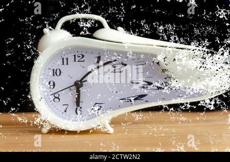 white alarm clock stands on a wooden table against a black wall. white alarm clock disintegrates into particles.concept of transience of time Stock Photo
