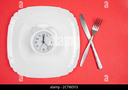 white alarm clock lies on a white plate with a fork and knife on a red background, top view Stock Photo