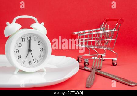 white alarm clock stands on a white plate on a red background with a fork and knife, a shopping cart close-up. concept of time for dinner, creative Stock Photo