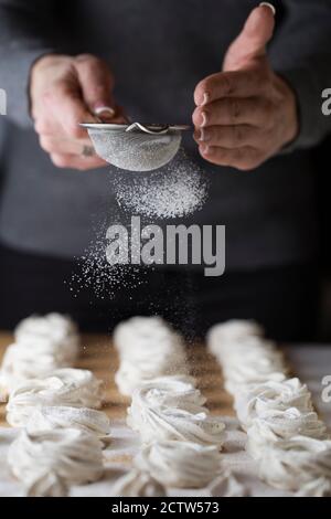 The process of making marshmallow. Close up hands of the chef with metal sieve sprinkling zephyr with Powdered sugar at pastry shop kitchen. confectio Stock Photo
