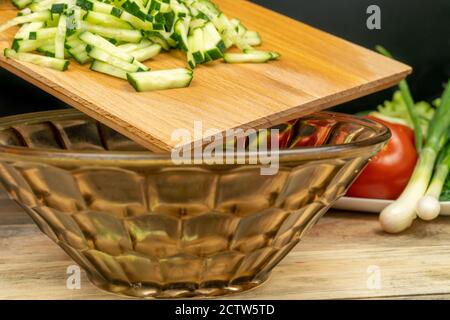 Sliced cucumbers from a wooden Board are placed in a salad bowl close-up, in the background a plate with vegetables Stock Photo