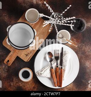 Crockery and cutlery on a dark background with copy space. White metal plates, bowls, cups and cutlery with wooden handles. Set of dishes Stock Photo