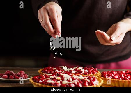 Beautiful freshly made cherry tart decorated with almond chips. Food photography. Process of making tart. Close up hands of the chef sprinkling cake w Stock Photo