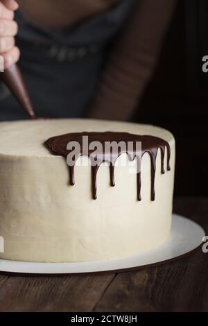 A confectioner squeezes liquid chocolate from a pastry bag on to a white cream biscuit cake. The concept of homemade pastry, cooking cakes. Stock Photo
