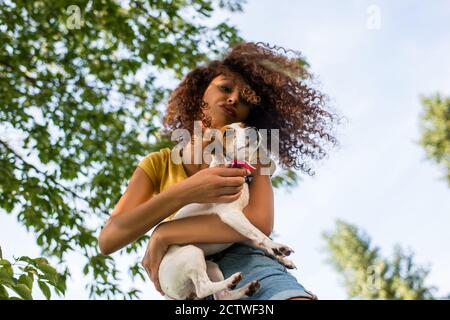 low angle view of curly woman holding jack russell terrier dog and making duck face against blue sky and branches Stock Photo