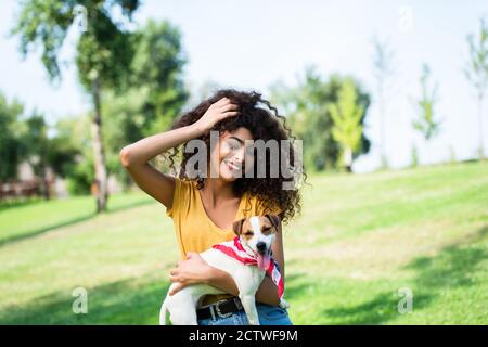 joyful woman touching curly hair while holding jack russell terrier dog in park Stock Photo