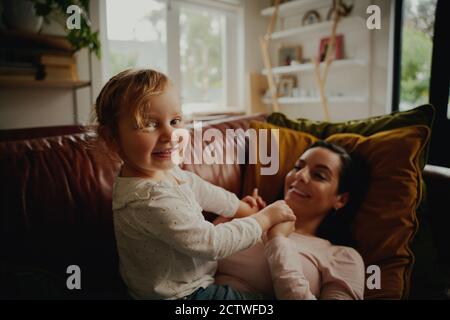 Portrait of excited little child playing with mother lying on couch at home during covid-19 lockdown