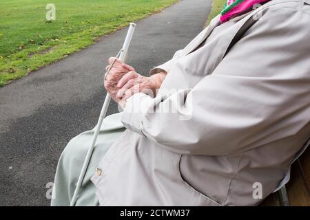 Ninety year old visually impaired lady with white stick sitting on bench in public park. England, UK Stock Photo