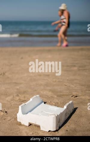 Plastic, white, styrofoam container left on sandy beach with walking people. Stock Photo