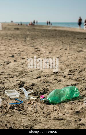 Green, plastic bottle, cotton swabs, cap, box and cigarettes left on a sandy beach with walking people in background. Stock Photo