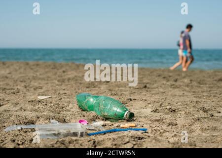 Green, plastic bottle, cotton swabs, cap, box and cigarettes left on a sandy beach with walking people in background. Stock Photo