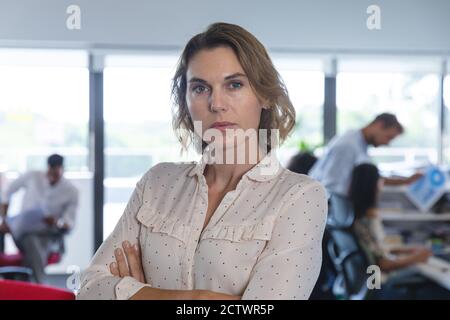 Portrait of confident Caucasian female business creative with arms crossed looking to camera, colleagues working in the background. Creative business Stock Photo