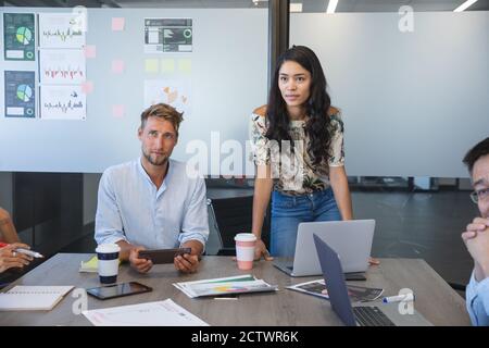 Asian businesswoman standing to address colleagues during a team meeting, a Caucasian male colleague sitting beside her listening. Creative business p Stock Photo