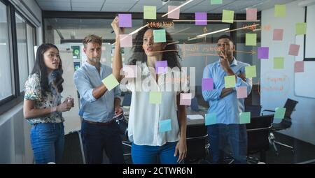 Multi-ethnic male and female business team brainstorming together, a mixed race woman writing on sticky notes stuck to a glass wall. Creative business Stock Photo