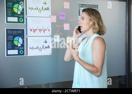 Smart casually dressed Caucasian businesswoman talking on smartphone, a board with charts and information on it in the background. Creative business p Stock Photo