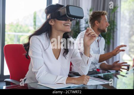 Asian businesswoman sitting at desk using VR headset and smiling, with male colleague discussing in the background. Creative business professionals wo Stock Photo