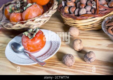 A persimmon served on a saucer with spoonfuls. In the background wicker baskets with other persimmons and dried fruit (walnuts and hazelnuts) Stock Photo