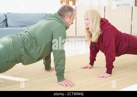 active mature couple doing plank exercise and looking each other in living room at home. Stock Photo