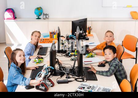 Selective focus of multicultural schoolkids looking at camera near robots and computers in classroom Stock Photo