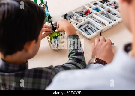Back view of teacher sitting near asian schoolboy modeling robot at table in stem school Stock Photo