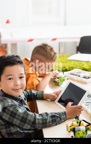 Selective focus of asian schoolboy holding digital tablet near friend modeling robot in stem school Stock Photo