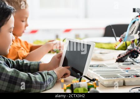 Selective focus of asian schoolboy using digital tablet near friend and robots on table in school Stock Photo