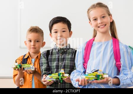 Selective focus of multiethnic pupils holding robots and looking at camera in stem school Stock Photo