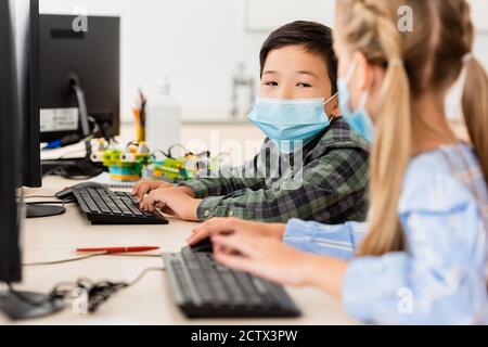 Selective focus of asian schoolboy in medical mask using computer near friend in classroom Stock Photo