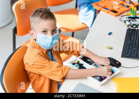 Schoolboy in medical mask holding building blocks near computer in classroom Stock Photo