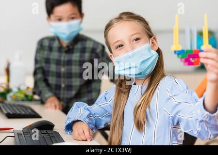 Selective focus of schoolgirl in medical mask holding building blocks near asian classmate in school Stock Photo