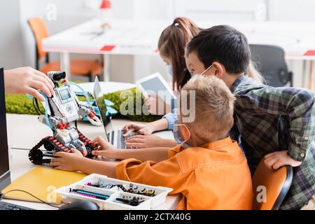 Selective focus of teacher holding robot near multiethnic pupils in medical masks and gadgets in classroom Stock Photo