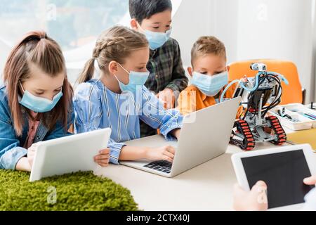 Selective focus of multiethnic children in medical masks using digital tablet and laptop near teacher and robot in school Stock Photo