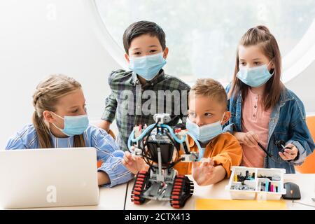 Selective focus of multiethnic classmates in medical masks constructing robot near laptop in classroom Stock Photo