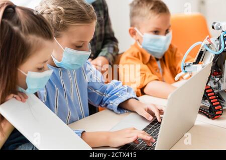 Selective focus of kids in medical masks using laptop and digital tablet near robot in school Stock Photo
