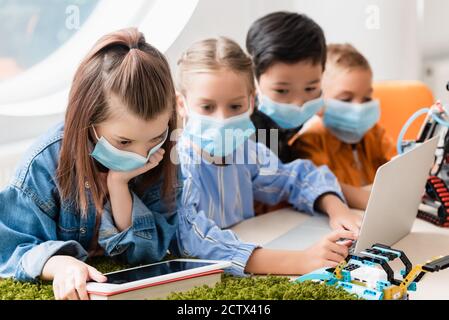 Selective focus of multiethnic schoolkids in medical masks using gadgets while programming robot in stem school Stock Photo