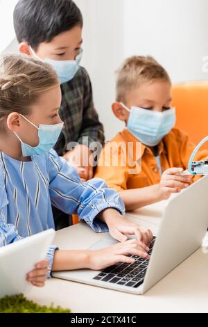 Selective focus of multicultural children in medical masks using gadgets while programming robot in stem school Stock Photo